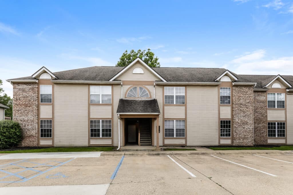 Two-story apartment building with tan siding, brick accents, and multiple windows, featuring a central entryway and nearby parking spaces, including accessible spots.
