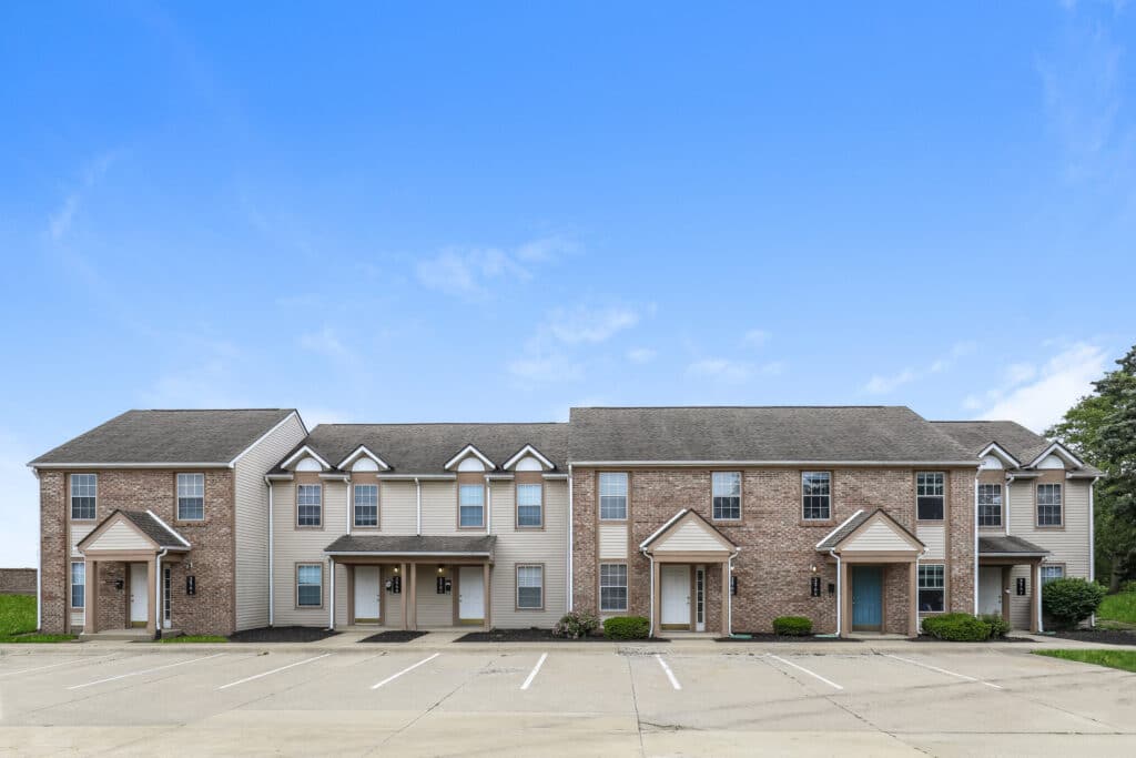 A row of two-story townhouses with brick and siding exteriors, multiple doors, windows, and empty parking spaces in front under a clear blue sky.
