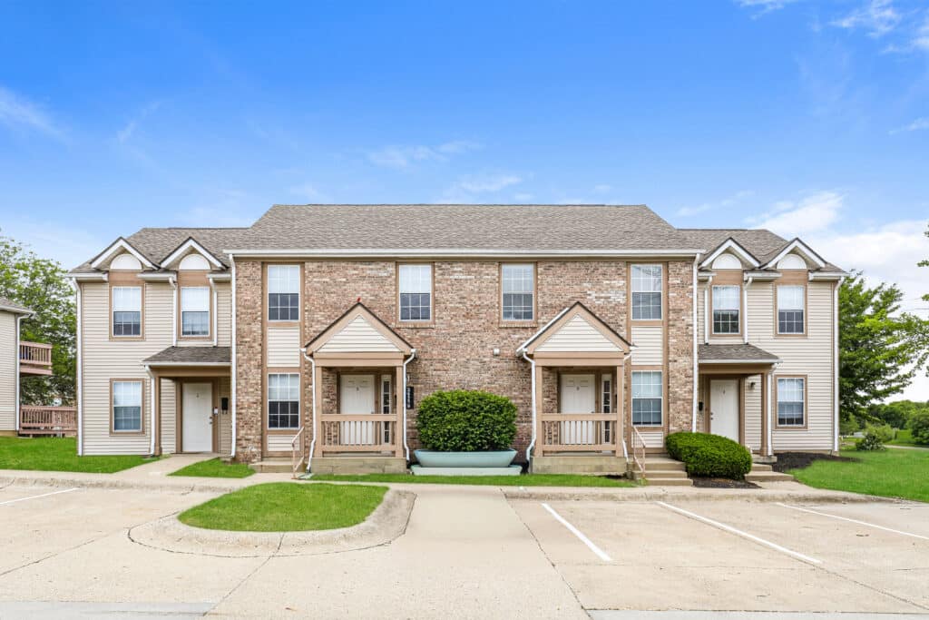 Brick and siding townhouse building with two entrances, shrubs, a green bench, and empty parking spaces in front under a clear blue sky.