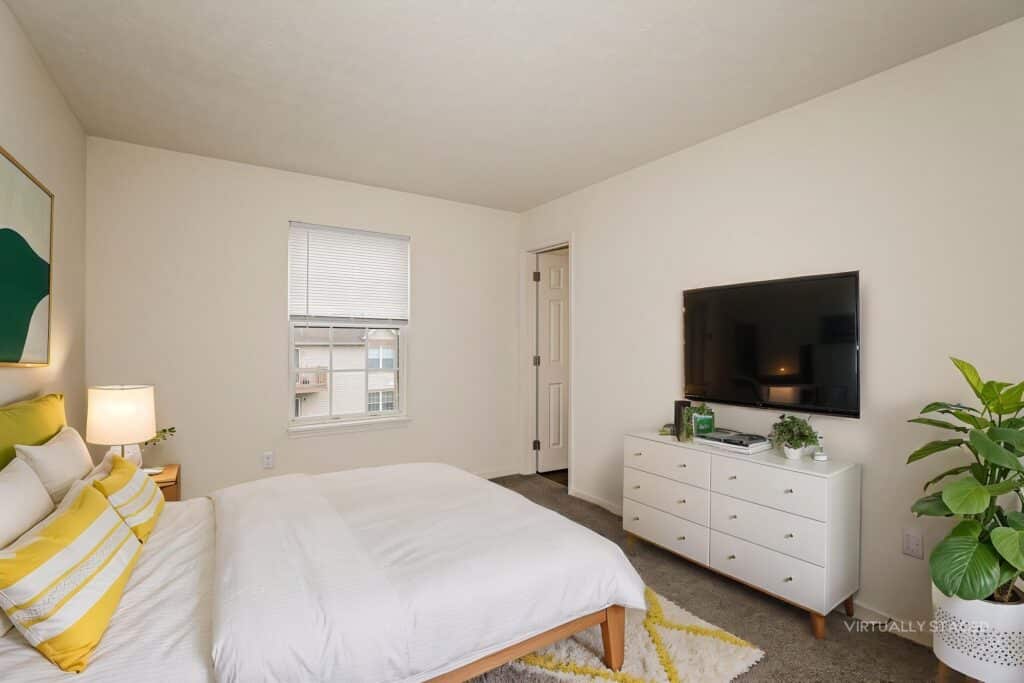 A bedroom with a bed, white dresser, large TV, window with blinds, indoor plant, and neutral decor.