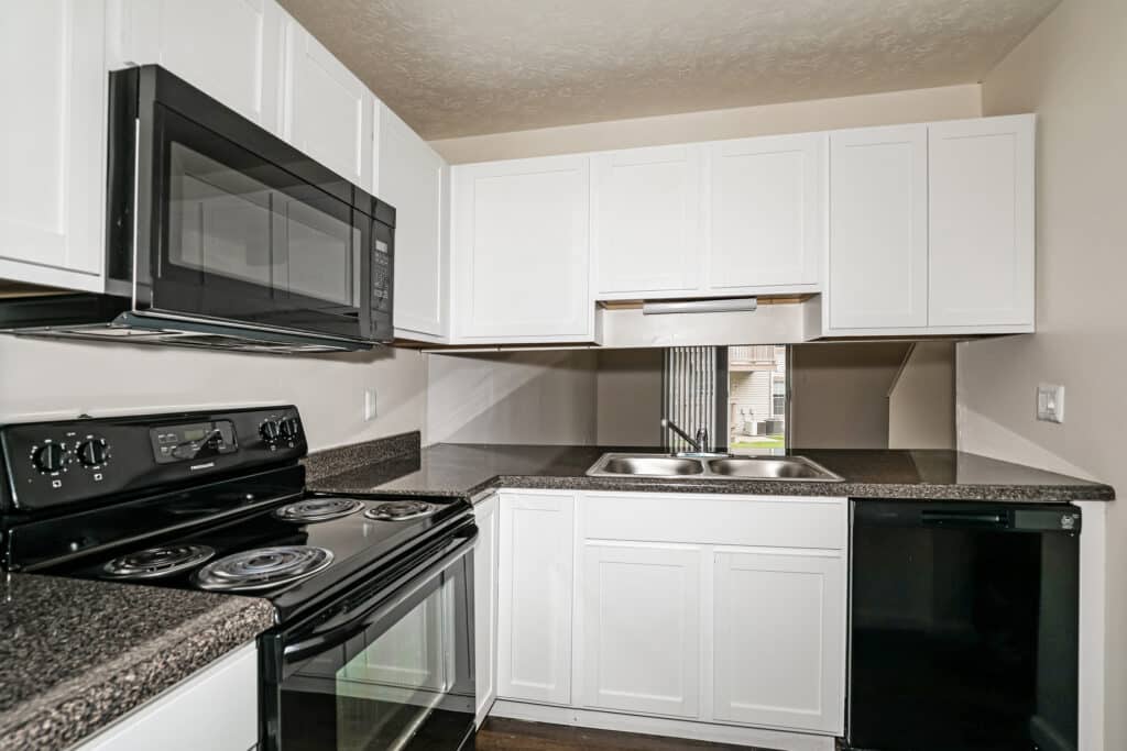 Modern kitchen with white cabinets, black appliances including stove, microwave, and dishwasher, and a stainless steel sink with a window above.