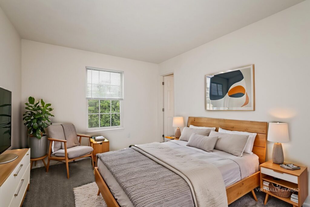 Modern bedroom with a wooden bed, neutral bedding, two nightstands with lamps, an armchair, a dresser, and a large window with blinds. Abstract art hangs above the bed.