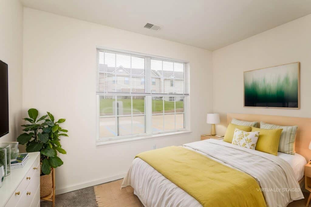 A bright bedroom with a double bed, green and yellow bedding, a dresser with a TV, a potted plant, two lamps, and a large window letting in natural light.