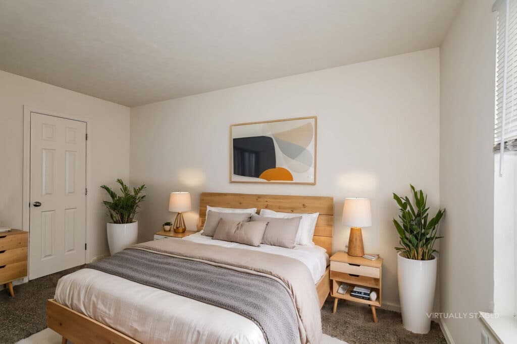 Minimalist bedroom with a wooden bed, neutral bedding, two bedside tables with lamps, abstract wall art, and potted plants on either side. Sunlight streams in through a window.