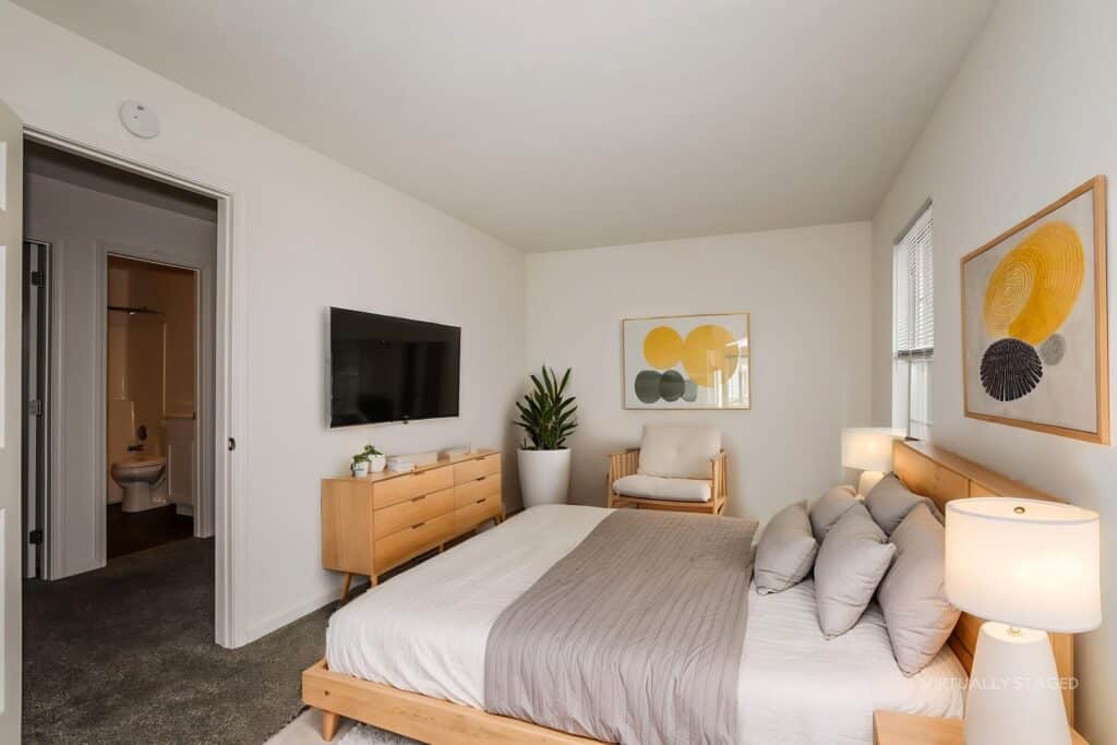 A modern bedroom with a wooden bed, gray bedding, two lamps, a dresser, a wall-mounted TV, a potted plant, artwork, and an armchair. A bathroom is visible through an open door.