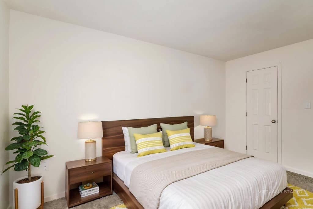Modern bedroom with a wooden bed, two bedside tables with lamps, green accent pillows, a potted plant, and a closed door in the background.