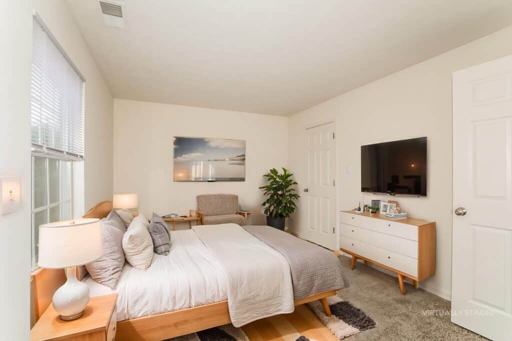 Bedroom with a bed, nightstand, armchair, dresser, TV, potted plant, and large window. Walls and bedding are light-colored; decor is minimal and modern. "Virtually staged" is noted.