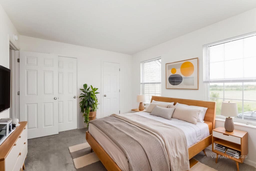 Bright bedroom with a wooden bed, neutral bedding, two windows, a plant, two bedside tables with lamps, and abstract artwork above the bed.