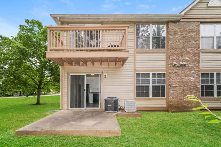Ground-level view of a beige and brick apartment exterior with a small patio, sliding glass door, balcony above, air conditioning unit, and green lawn.