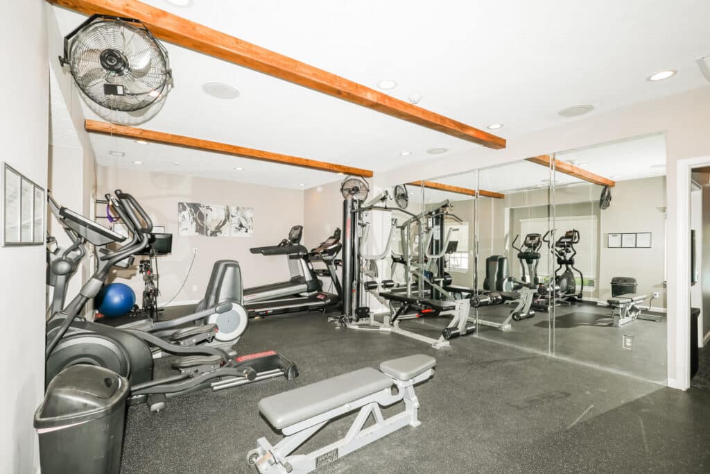 A home gym with various exercise machines, free weights, a bench, wall mirrors, and ceiling fans in a bright, well-lit room.
