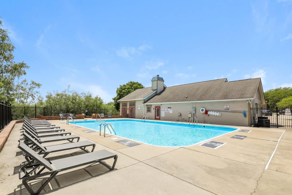 Outdoor swimming pool with clear water, surrounded by lounge chairs, and adjacent to a single-story clubhouse under a clear blue sky.