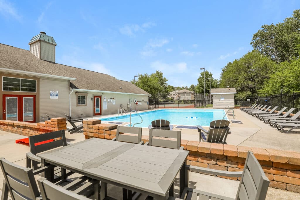 Outdoor swimming pool area with lounge chairs, patio tables, and a building with red-framed doors under a clear blue sky.