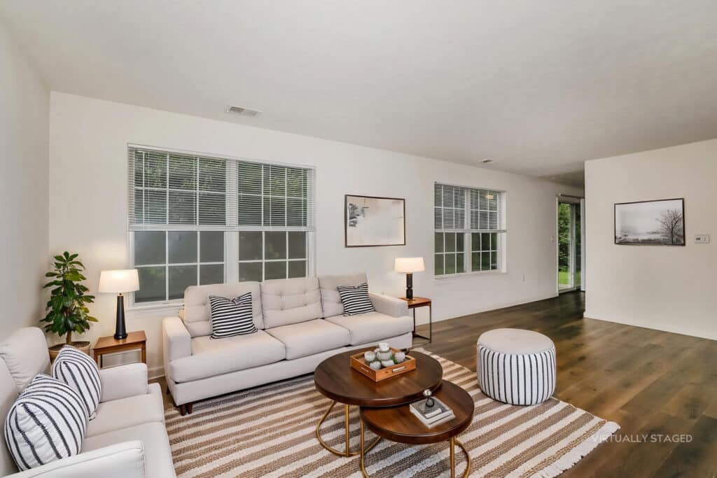 Living room with white sofa, armchair, striped pillows, two round wooden coffee tables, a pouf, striped rug, plants, and wall art. Large windows let in natural light; hardwood floors throughout.