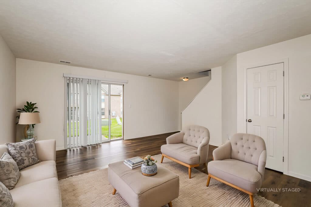 A minimally furnished living room with a sofa, two armchairs, a small table, and sliding glass doors leading to a grassy backyard; stairs and a closed door are visible.
