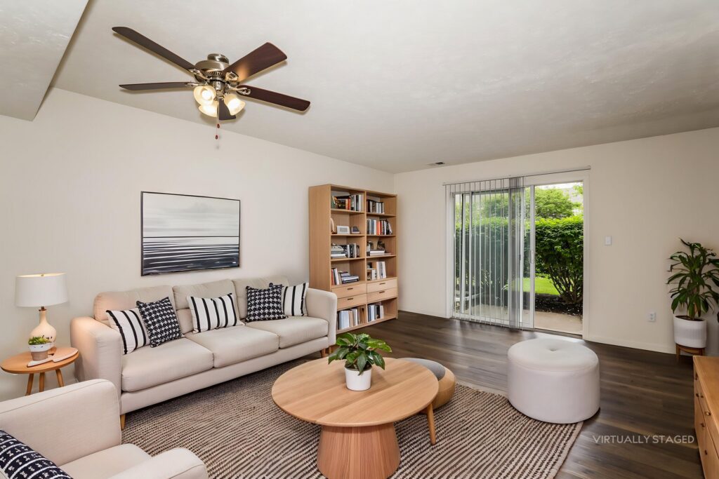 A living room with a ceiling fan, sofa, round coffee table, bookshelf, and sliding glass door leading to a garden.