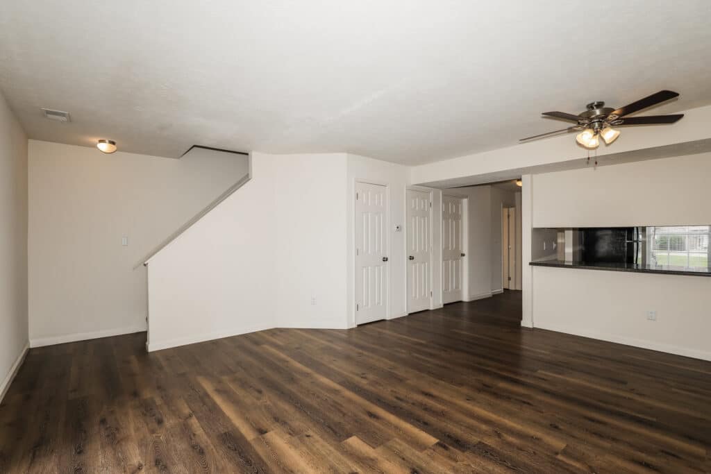 Empty living room with wood flooring, white walls, ceiling fan, staircase to the left, multiple white doors, and a kitchen pass-through window on the right.