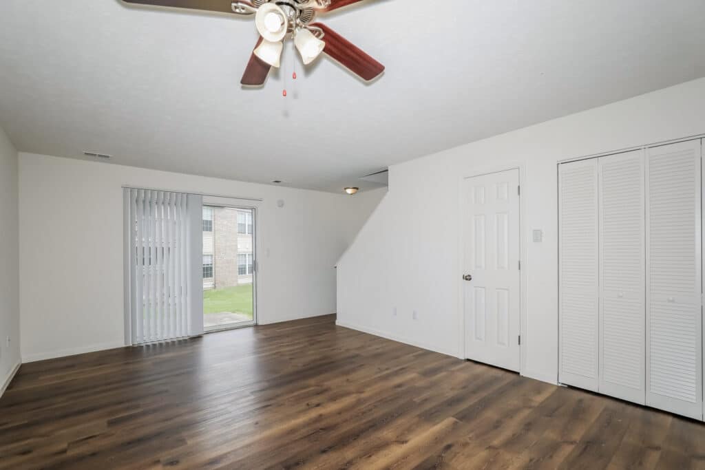 Unfurnished living room with wood floors, white walls, ceiling fan, closet doors, stairway, and sliding glass door leading outside.