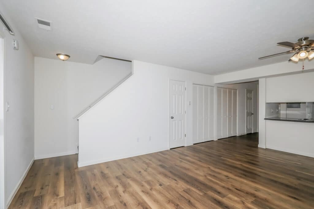 Empty room with wood flooring, white walls, a staircase, ceiling fan, closet doors, and a pass-through window to the kitchen.