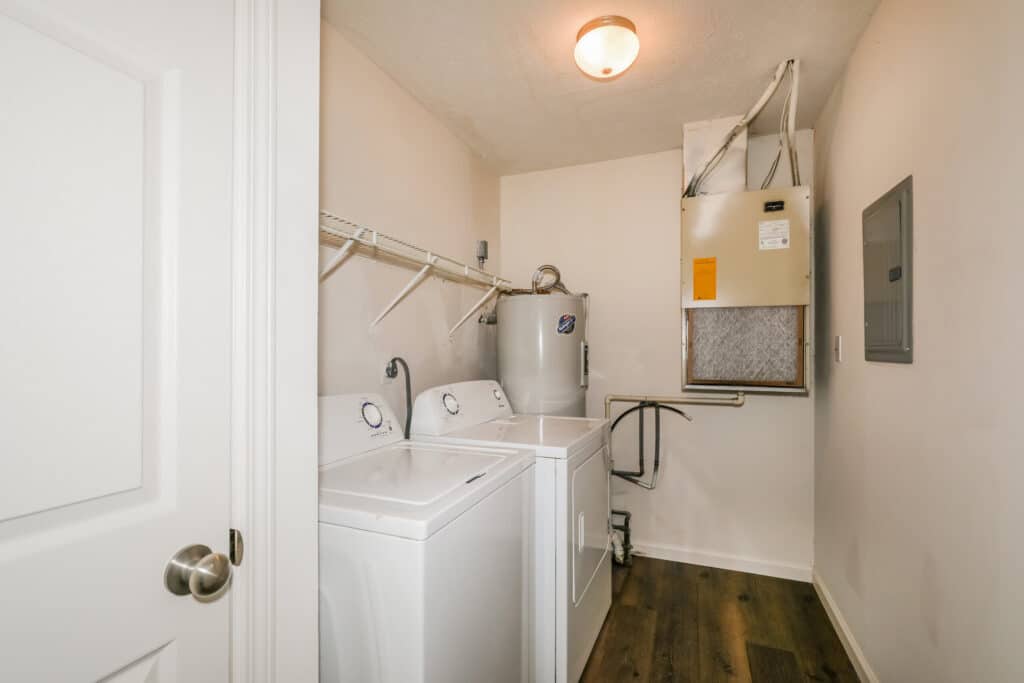 Laundry room with a white washer and dryer, water heater, HVAC unit, overhead wire shelf, and wood flooring.