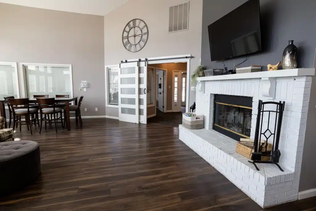 Modern living room with a white brick fireplace, wall-mounted TV, dining table, large wall clock, sliding glass doors, and wooden floors.