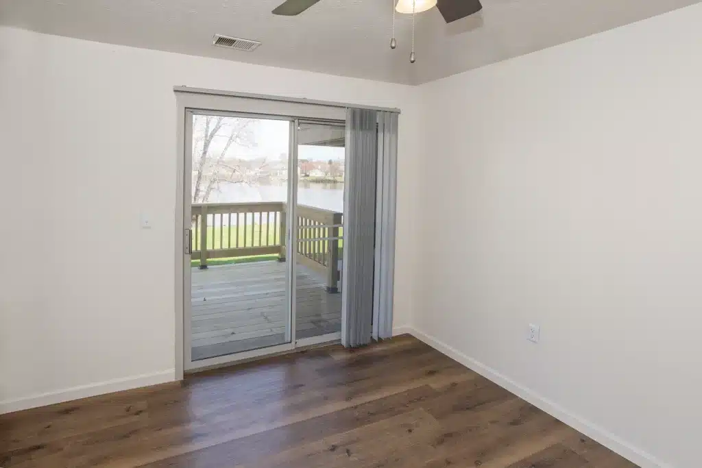 Empty room with light-colored walls, wood flooring, ceiling fan, and a sliding glass door leading to a covered patio with a view of a lake and grassy area outside.