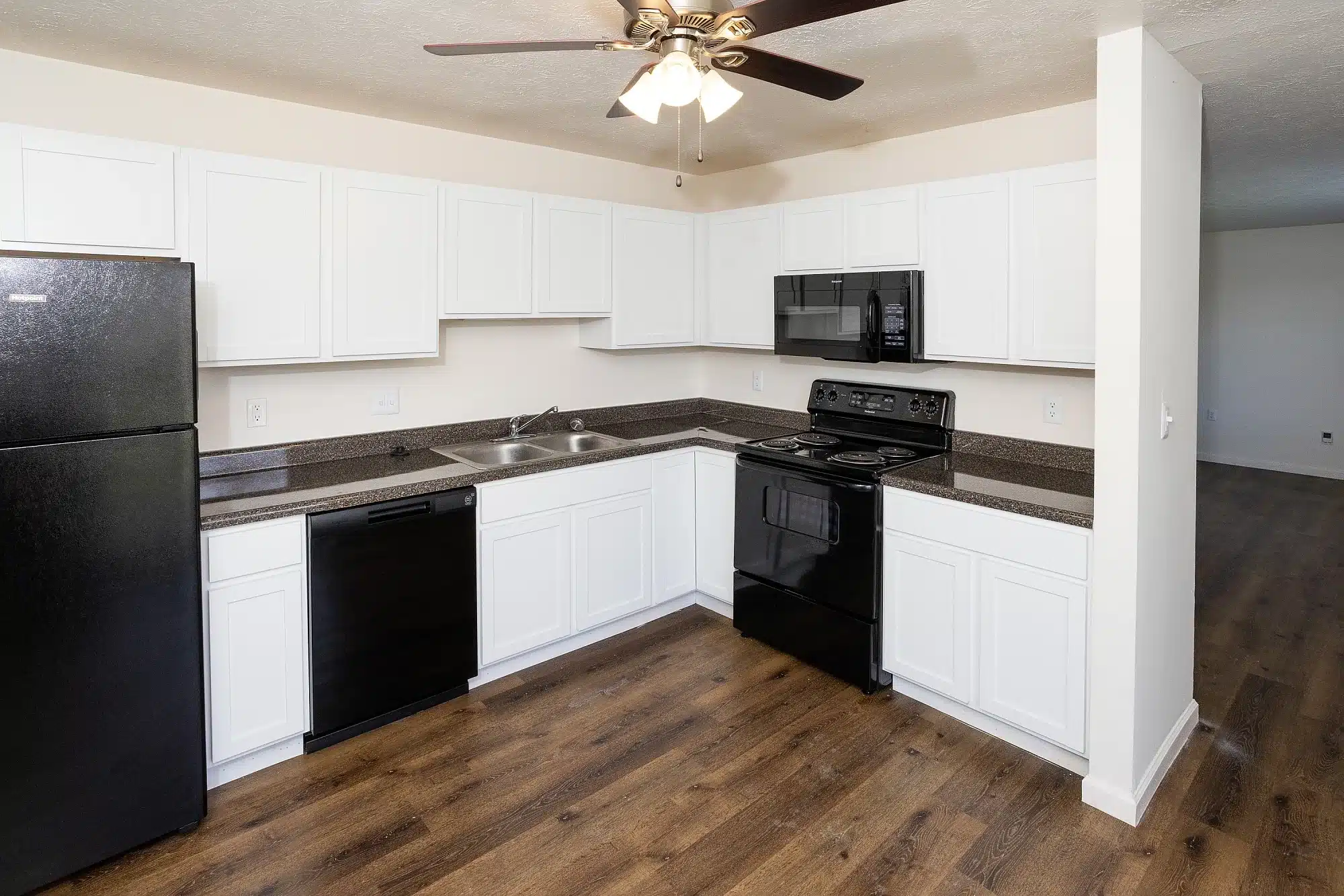 Modern kitchen with white cabinets, black appliances including fridge, stove, microwave, and dishwasher, brown countertops, and wood-style flooring. Ceiling fan with lights above.