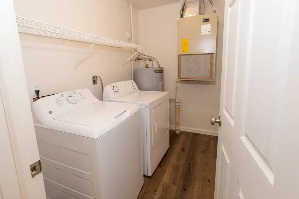 A laundry room with a washing machine, dryer, water heater, HVAC unit, wire shelf above, and wood-look flooring.