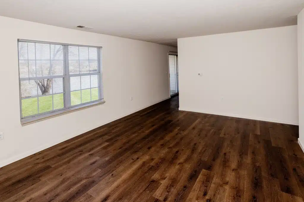 Empty room with white walls, large window with blinds, and dark wood flooring. Natural light enters from the window, and the room appears clean and unfurnished.