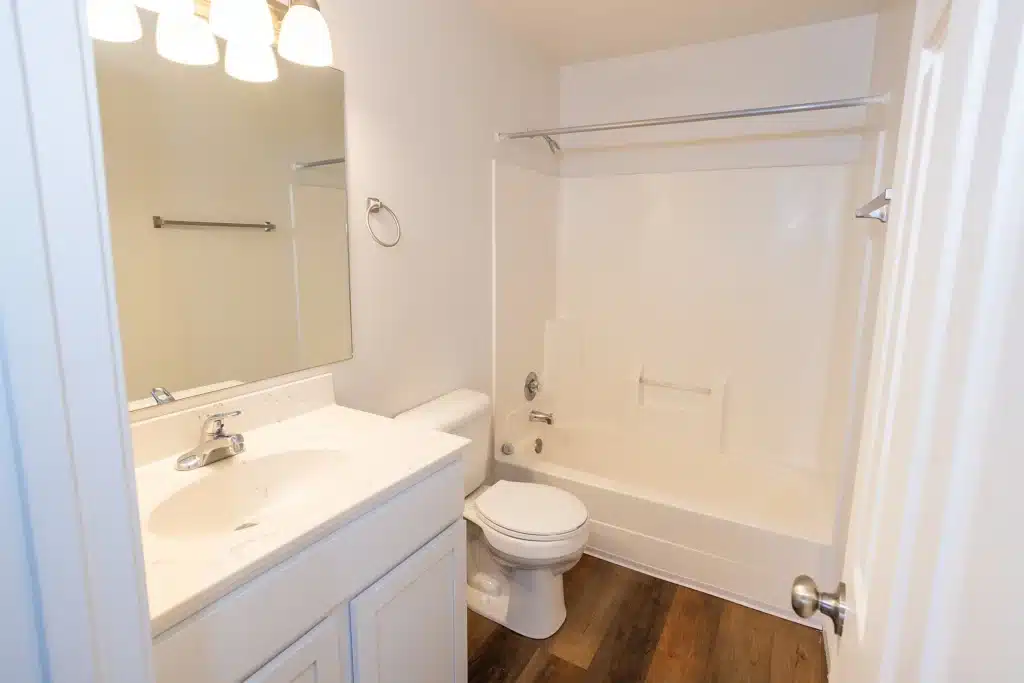 White bathroom with a sink, mirror, toilet, and bathtub with shower, featuring wood-look flooring and simple fixtures.