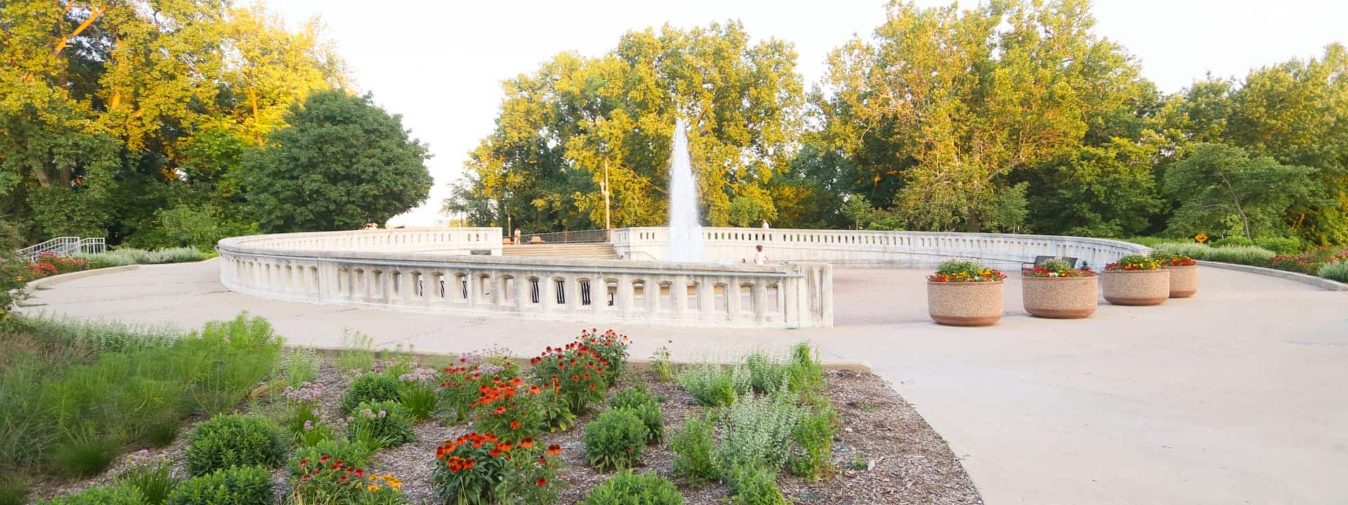A circular concrete fountain surrounded by a white railing, large planters, flower beds, and trees in the background on a sunny day.