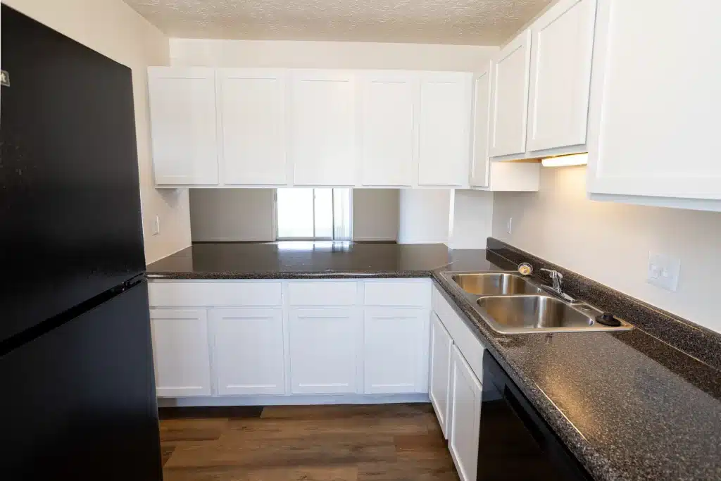 A modern kitchen with white cabinets, black appliances, a double sink, and dark countertops, featuring wood-style flooring and a small window above the counter.