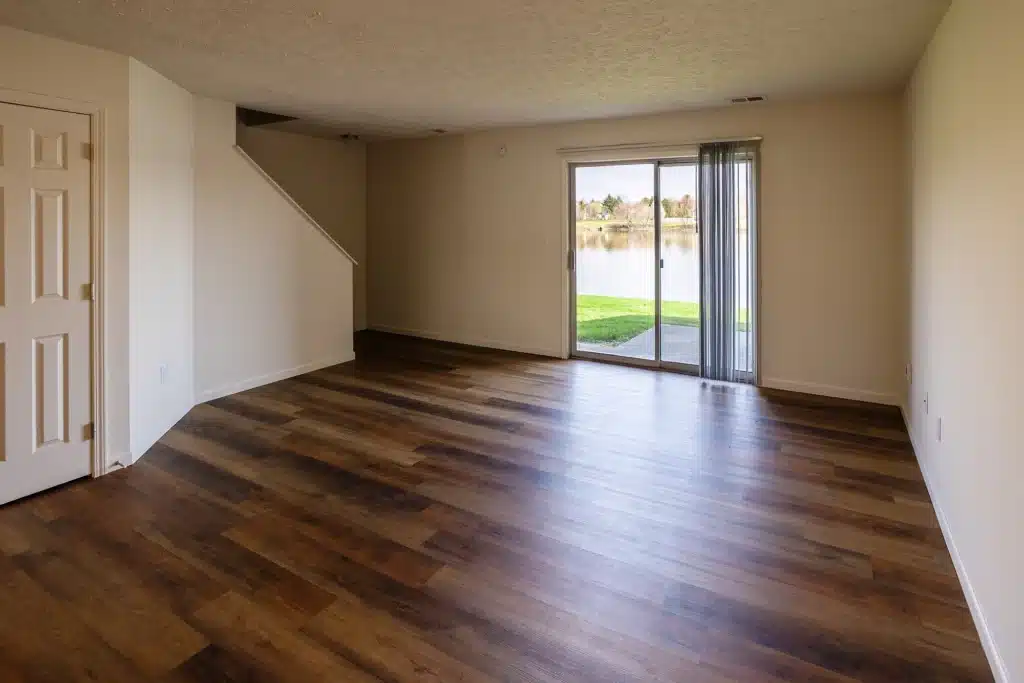 Empty living room with wood-style flooring, beige walls, a staircase, and sliding glass doors leading to a grassy area by a lake.