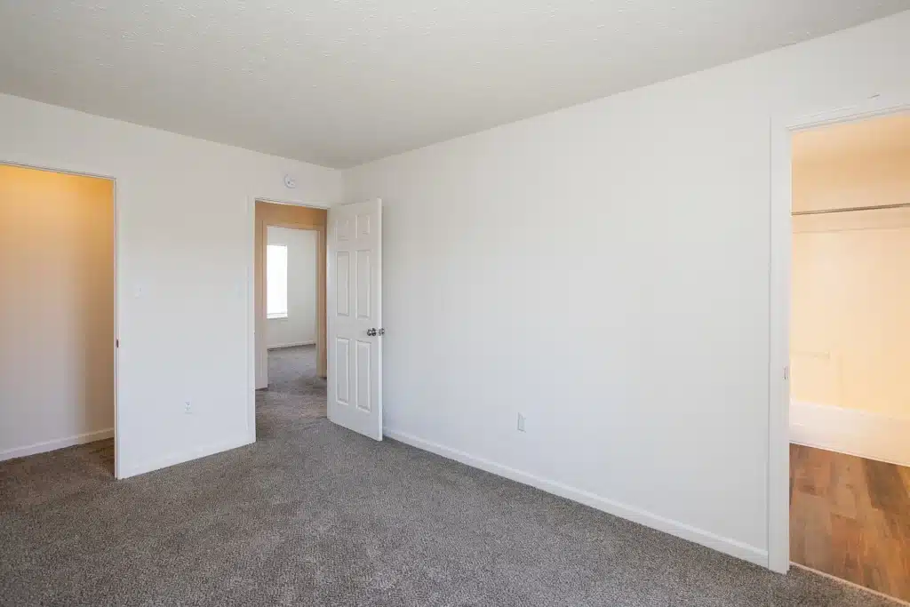 Empty bedroom with beige carpet, white walls, open door to hallway, doorway to bathroom with tub, and a closet with wooden floor visible.