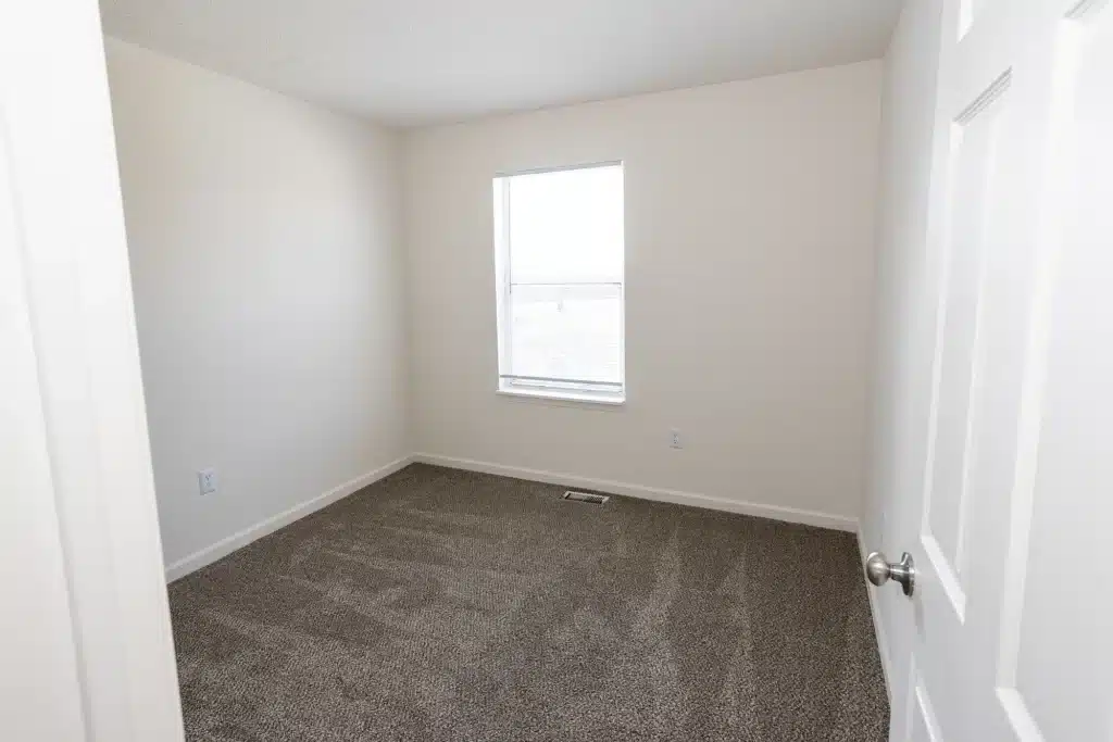 An empty room with beige walls, a single window, and gray carpet, viewed from the doorway.