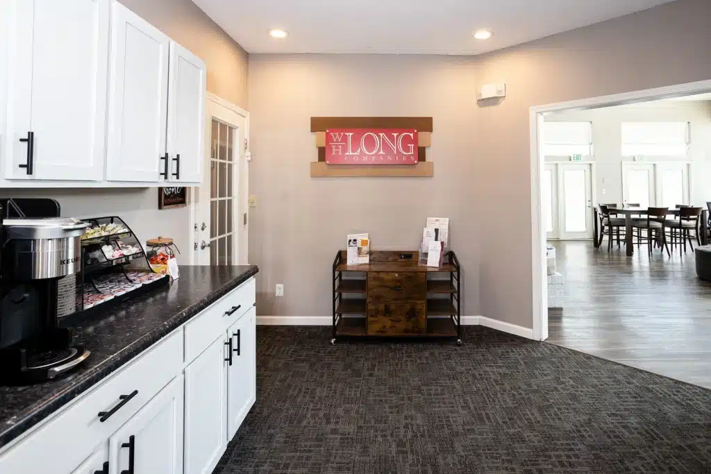 Office kitchenette area with white cabinets, coffee station, and brochures on a cart; adjoining room visible through wide doorway with tables and chairs.
