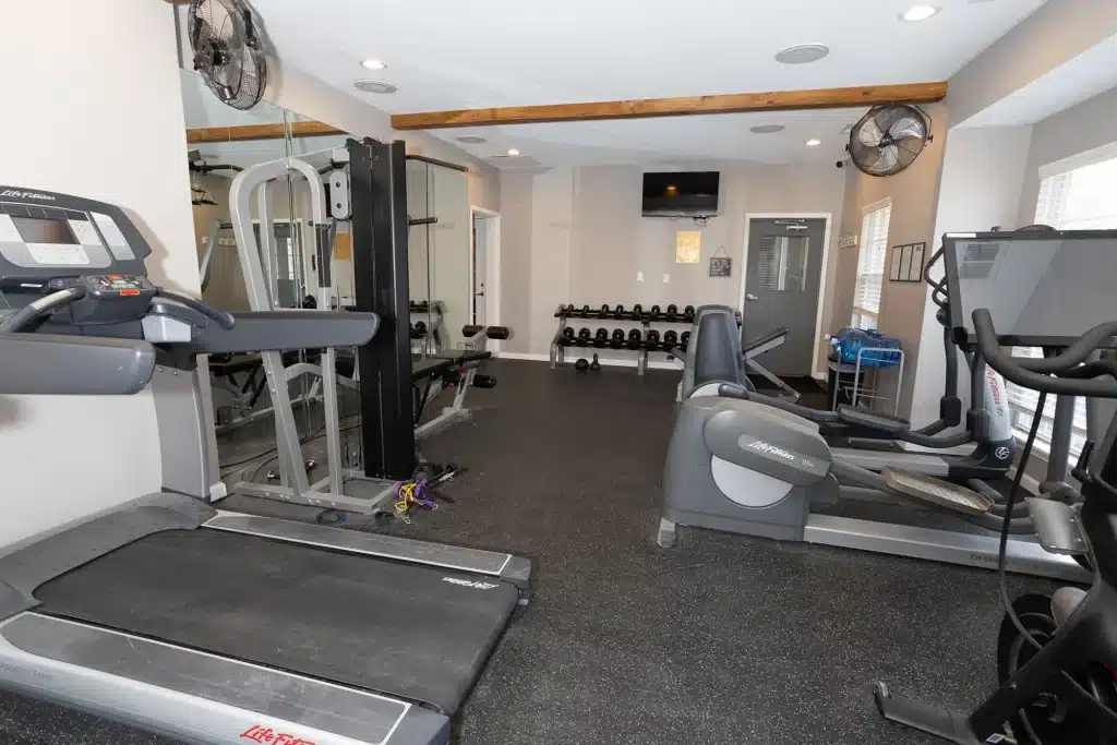 Small gym with treadmills, elliptical machines, weight machines, free weights, wall mirrors, fans, and a TV mounted on the wall.