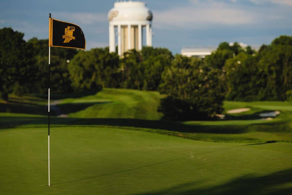 A Purdue University golf flag stands on a green, with a white water tower and trees in the background under a partly cloudy sky.