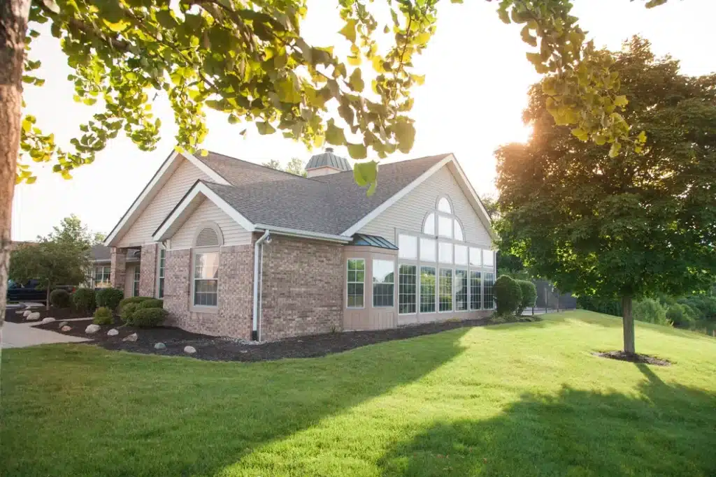 A single-story brick house with large windows sits on a well-maintained lawn, surrounded by trees and shrubs under bright sunlight.