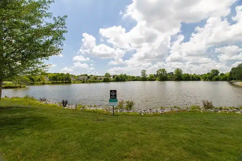 A pond surrounded by grass and trees under a partly cloudy sky, with a small sign posted near the water’s edge.